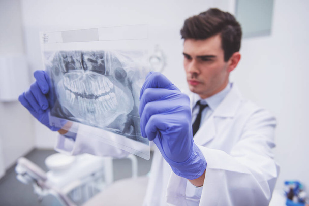 Молодий лікар стоматолог в захисних рукавичках, дивлячись на рентгенівський знімок щелепи людини
. - Фото, зображення