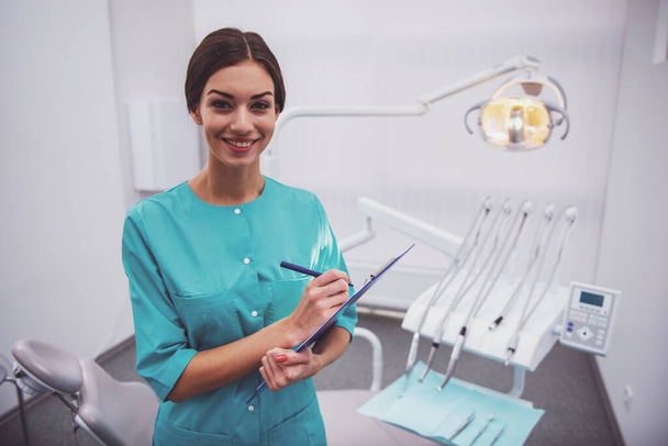Портрет молодой женщины-стоматолога, запись в документе, на заднем плане стоматологическая клиника
 - Фото, изображение