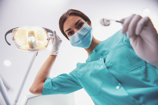Young woman dentist in protection gloves and a mask, keeps dental instrument looking at the camera. - Fotoğraf, Görsel