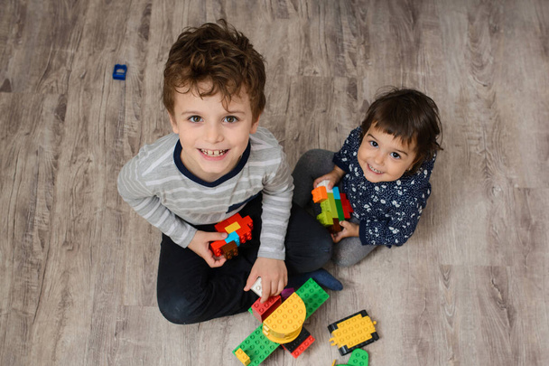 Top view of two happy children brother and his sister they are playing with plastic toy building bricks, smile and looking to the camera. They are sitting on the wood floor - Foto, Bild