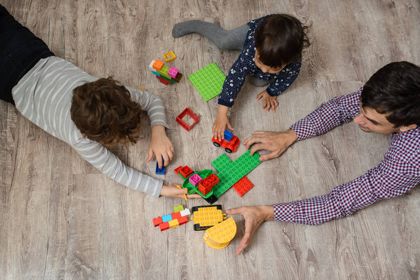 Bovenaanzicht van vader met zijn twee kinderen broer en zus spelen ze met plastic speelgoed bouwstenen. Ze liggen op de houten vloer. - Foto, afbeelding