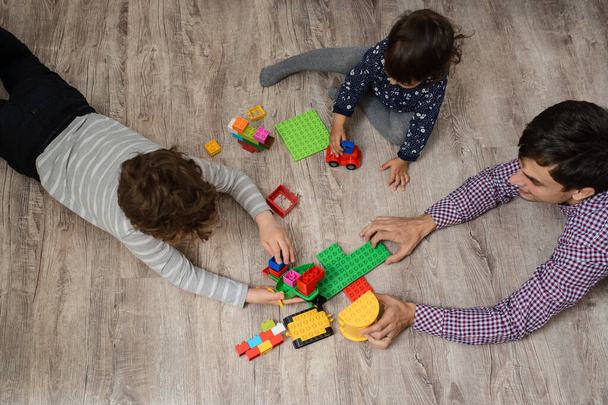 Vista dall'alto del padre con i suoi due figli fratello e sorella stanno giocando con mattoni da costruzione in plastica giocattolo. Sono sdraiati sul pavimento di legno - Foto, immagini