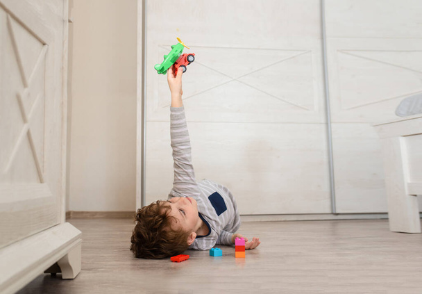 Szczęśliwy chłopiec leży na drewnianej podłodze i bawi się zabawką i plastikowymi cegłami. - Zdjęcie, obraz
