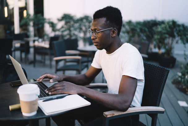Serious dark skinned male it developer typing on laptop computer making research and watching video on cafe terrace, handsome man in eyewear watching webinar for online learning course - Фото, зображення