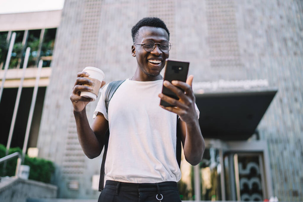 Szczęśliwy ciemnoskóry mężczyzna tysiąclecia w casual nosić ciesząc się oglądanie filmów z vlogów na smartfonie na świeżym powietrzu, uśmiechnięty Afroamerykanin człowiek trzyma filiżankę kawy i poszukiwanie dobrych wiadomości na telefon komórkowy - Zdjęcie, obraz