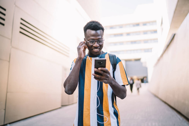 Happy African American blogger in classic eyewear and electronic earphones choosing music playlist on modern smartphone technology, millennial hipster guy enjoying positive song and online chatting - Foto, imagen