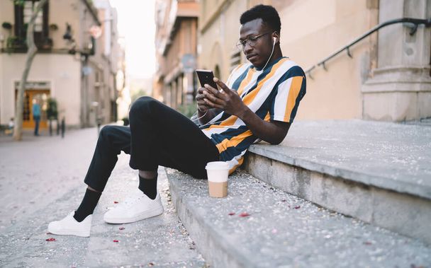 African American hipster guy in eyewear using cellular app for online phoning during travel vacations, young male blogger in modern headphones checking received email and read network publication - 写真・画像