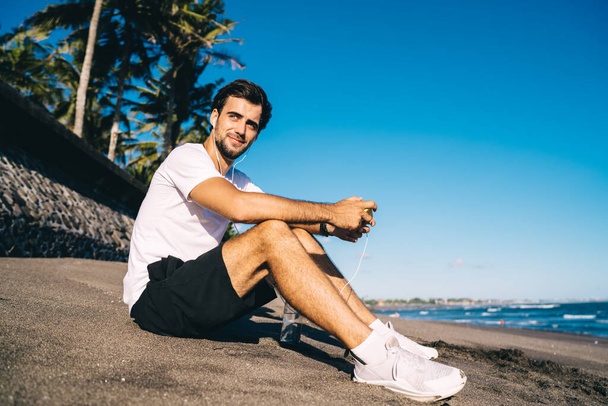 Low angle side view of slim cheerful guy in sportswear and sneakers listening to music in earphones while sitting on sand coast near ocean under serene sky and looking away - Fotoğraf, Görsel