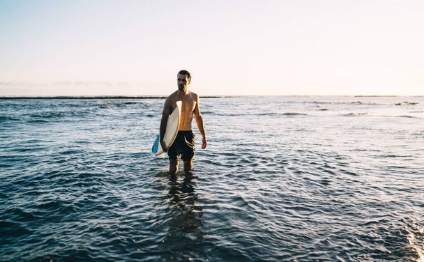 Nyugodt, fiatal, félmeztelen, izmos férfi sportoló fürdőruhában, egyedül áll az óceán hullámzó hullámaiban, és naplementekor szörfdeszkával a kezében néz félre. - Fotó, kép
