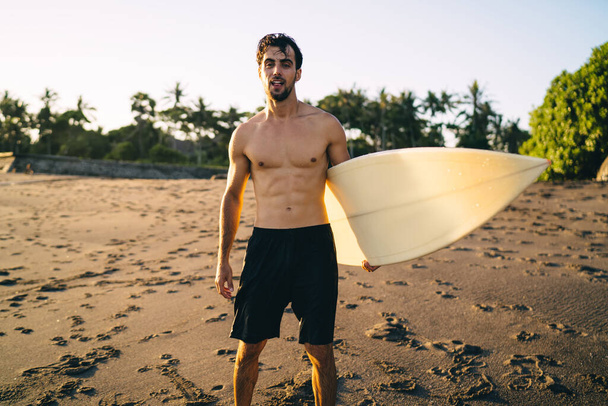 Confident young bearded male athlete with naked torso standing on sandy beach with surfboard in hand and looking at camera during golden hour in tropical resort - Fotografie, Obrázek