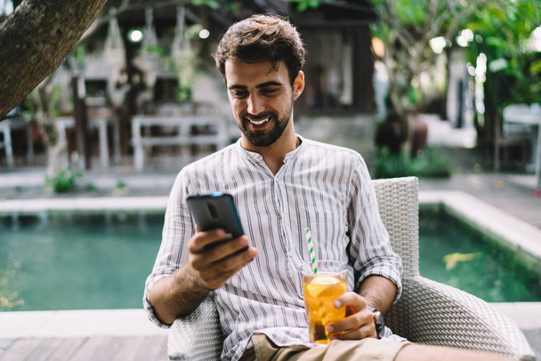 Slim adult glad guy text messaging on cellphone while sitting on comfortable chair with glass of colorful ice tea and straw near swimming pool in cafe - Фото, зображення