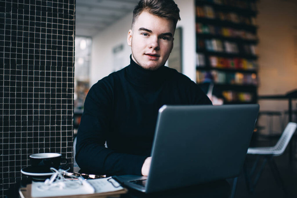 Positive young man in black turtleneck using laptop for surfing internet and looking at camera with half smile while having break in empty library - Zdjęcie, obraz