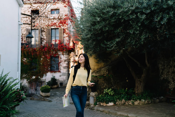 Positive ethnic female in casual clothes and backpack with toothy smile walking on street and holding map while looking away - Fotografie, Obrázek
