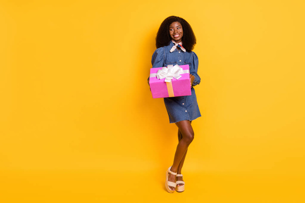 Full body portrait of charming glad dark skin girl look empty space hold giftbox isolated on yellow color background - Zdjęcie, obraz