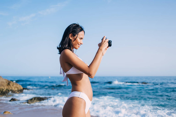 Side view of cheerful Latin woman in bikini swimsuit enjoying photo hobby while testing vintage camera and making images of ocean horizon, happy female swimmer in white bikini using retro technology - Foto, Bild