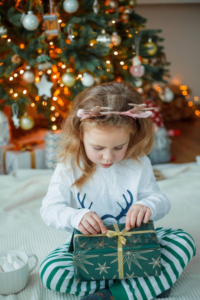 Suloinen 3-vuotias tyttö pyjamassa avaa lahjapakkauksen joulukuusen lähellä. Uudenvuodenvalot. Talvilomat. Kodikas koti. - Valokuva, kuva