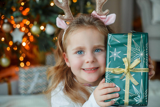 Adorable 3 year old girl in pajamas opens gift box near the Christmas tree. New Year's lights. Winter holidays. Cozy home. - Foto, imagen