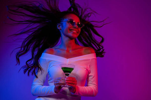 Fénykép lenyűgözött fiatal barna hölgy ital alkohol megjelenés hirdetés visel fehér ruha szemüveget elszigetelt felett neon színes háttér - Fotó, kép
