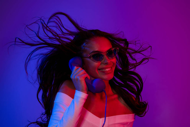 Photo of sexy young brunette lady talk telephone wearing white dress spectacles isolated over neon color background - Foto, Imagen