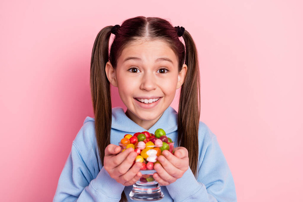 Photo portrait of happy schoolgirl keeping glass with sweet candies isolated on pastel pink color background - 写真・画像