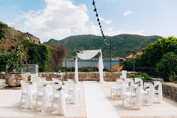 Белые стулья украшены тканевым стендом перед свадебной аркой в саду у моря - Фото, изображение
