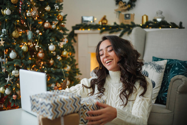 明るいニットのセーターを着た幸せな若い暗い髪の巻き女性は、クリスマスツリーの近くに贈り物を準備します。スタイリッシュなモデルは、冬の休暇中にオンライン購入を行います。新年。居心地の良い家. - 写真・画像