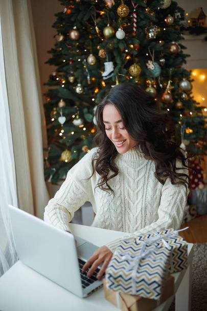 Happy young dark-haired curly woman in a light knitted sweater prepares gifts near the Christmas tree. Stylish model makes online purchases during the winter holidays. New Year. Cozy home. - Zdjęcie, obraz