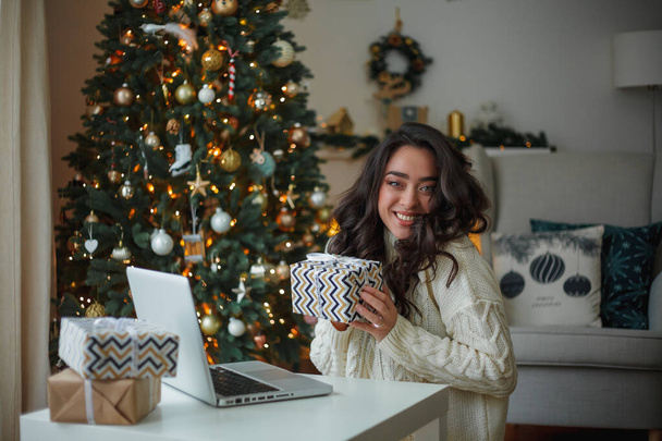 明るいニットのセーターを着た幸せな若い暗い髪の巻き女性は、クリスマスツリーの近くに贈り物を準備します。スタイリッシュなモデルは、冬の休暇中にオンライン購入を行います。新年。居心地の良い家. - 写真・画像