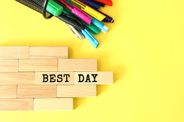 Blocchi di legno impilati accanto a penne e matite su uno sfondo giallo. BEST DAY testo su un blocco di legno. - Foto, immagini