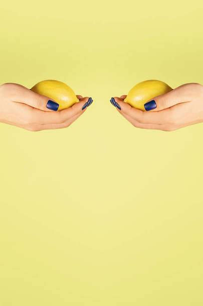 Two open female hands with blue nails holding fresh lemons opposite each other isolated on yellow. Vertical. Copy space - Zdjęcie, obraz