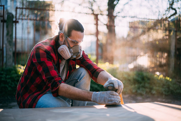 A male carpenter works with a wood grinder. Professional equipment and protective equipment. Young professional sanding wood table top outdoors. - Foto, Imagem