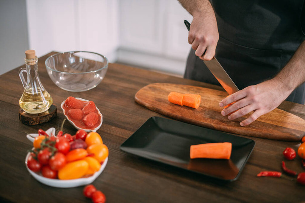 Ένας αρσενικός μάγειρας ετοιμάζει λαχανικά στο σπίτι στην κουζίνα. Κοντινό πλάνο των χεριών με ένα μαχαίρι και λαχανικά. Νεαρός με ποδιά στην κουζίνα του σπιτιού στο τραπέζι με υλικά για βραστό λαχανικών ή σαλάτα - Φωτογραφία, εικόνα