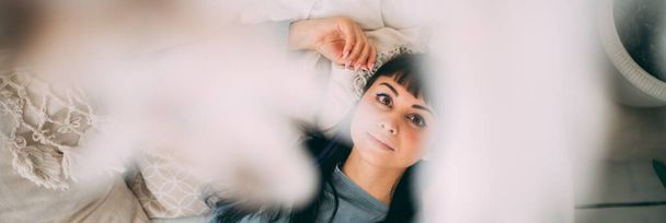 Retrato de una joven soñadora en la cama en pijama azul en un acogedor dormitorio. Hermosa mujer morena en ropa cómoda para el hogar está descansando en una cama grande en un interior de estilo boho - Foto, Imagen