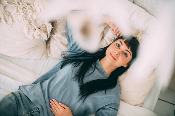 Portrait d'une jeune fille rêveuse sur le lit en pyjama bleu dans une chambre confortable. Belle femme brune dans des vêtements confortables à la maison repose sur un grand lit dans un intérieur de style boho - Photo, image