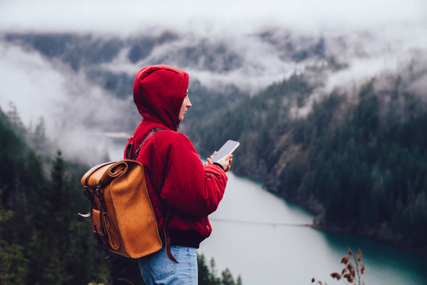 Side view of female tourist in warm clothes with hood and backpack messaging smartphone while exploring beautiful landscape with forest and river in USA - Foto, Imagem