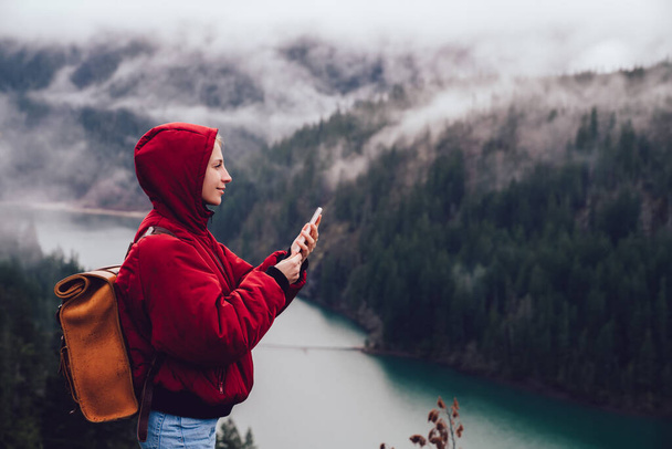 Side view of positive calm female in warm coat with hood and backpack messaging smartphone while discovering natural scenery with river and trees in fog - Photo, image