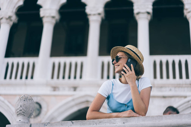 Low angle of trendy female explorer standing near white columns while leaning on railing and listening to partner on cellphone - Фото, изображение