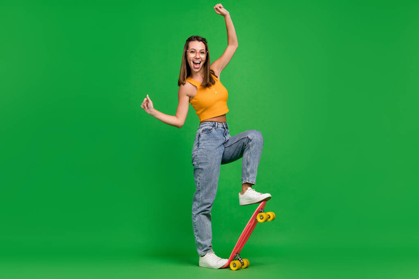 Full length photo of young joyful happy cheerful woman wear glasses ride longboard isolated on green color background - Φωτογραφία, εικόνα