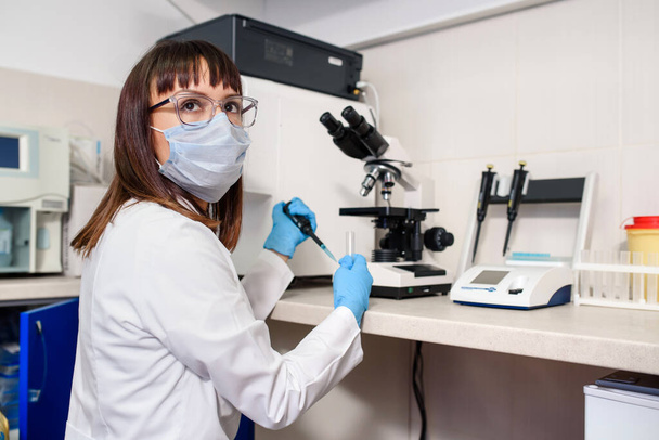 Young woman doctor laboratory assistant is using micropipette test tubes near a medical microscope and urine strip analyser. In the background a fully-automated diagnostic chemistry system for immunological tests. The hospital laboratories research p - Foto, Bild