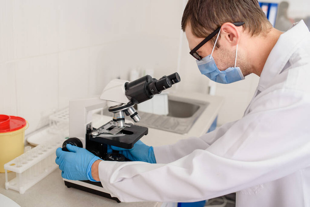 В больничных лабораториях, в процессе работы, молодой человек-врач смотрит в медицинский микроскоп на анализ крови. - Фото, изображение