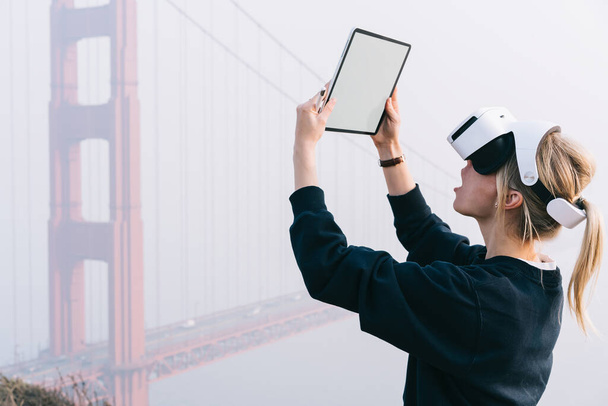 Side view of blond astonished female in casual clothing using tablet with blank screen and VR goggles in San Francisco near famous bridge - Фото, изображение