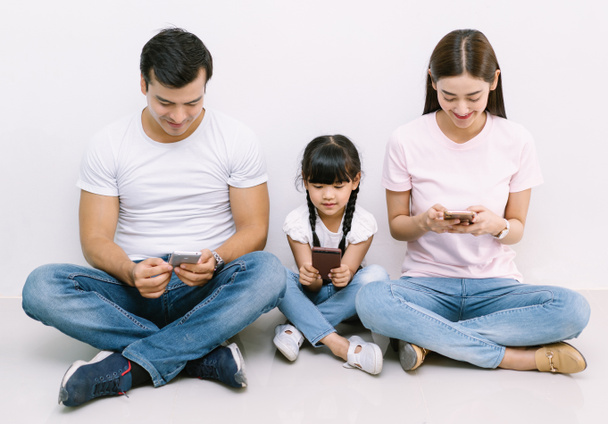 Happy technology addicted Asian family parents and kid holding smart phones having electronic devices sit on floor white background. Gadgets dependence overuse, Social media addiction concept - Foto, Bild