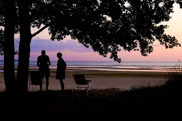 a man and a woman are standing on the shore of a lake in the setting sun, standing near camping chairs. two people on the background of the ocean in the evening - Fotoğraf, Görsel