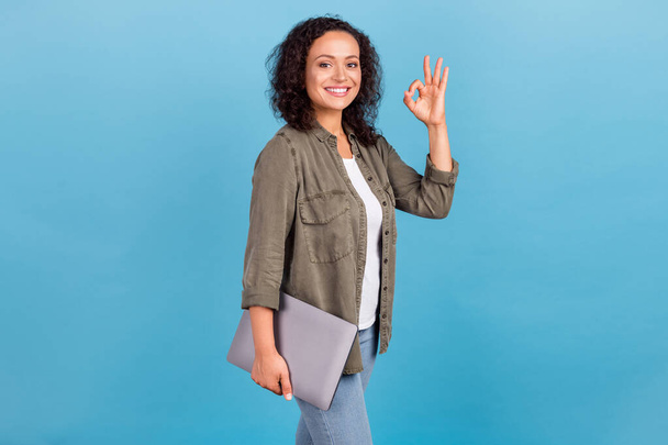 Portrait of attractive cheerful wavy-haired girl carrying laptop showing ok-sign isolated over bright blue color background - Foto, Imagem