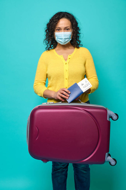 Young woman in protective medical mask, wearing bright yellow sweater posing with suitcase, passport and boarding pass in her hands. Travel, air flight concept on blue background, copy ad space - Photo, Image