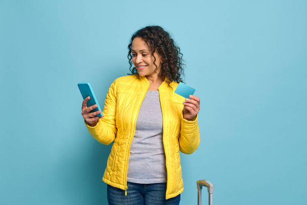 Jolie femme souriante portant une veste jaune, souriant tenant une carte bleue de crédit ou de réduction avec espace de copie et glisser, naviguer sur les sites Web, réserver sur un téléphone mobile, isolé sur fond bleu - Photo, image
