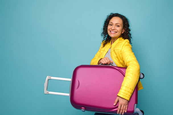 Charmante jonge vrouw reiziger in felgele jas, glimlachend met vrolijke tandenglimlach kijken naar de camera en houden haar koffer onder de armen, geïsoleerd op blauwe achtergrond met kopieerruimte - Foto, afbeelding
