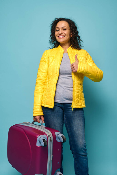 Beautiful delighted curly-haired woman in bright yellow jacket, holding her suitcase and showing thumb up, smiling toothy smile looking at camera, isolated over blue background with copy ad space - Foto, Bild