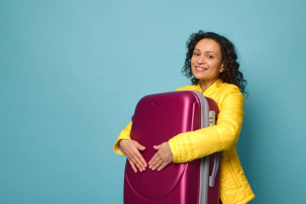 Beautiful woman in bright yellow jacket smiles toothy smile, posing with a colorful burgundy suitcase in the hands, isolated over blue background with copy ad space. Travel, tourism concept - Foto, Bild
