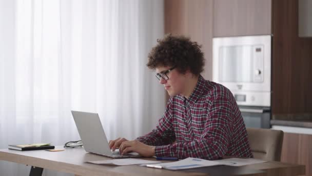 Un hombre rizado con una mirada seria trabaja en un ordenador portátil, sentado en una cocina moderna. Hombre joven freelancer estudiante utilizando el ordenador portátil que estudia en línea trabajando desde casa en Internet - Imágenes, Vídeo